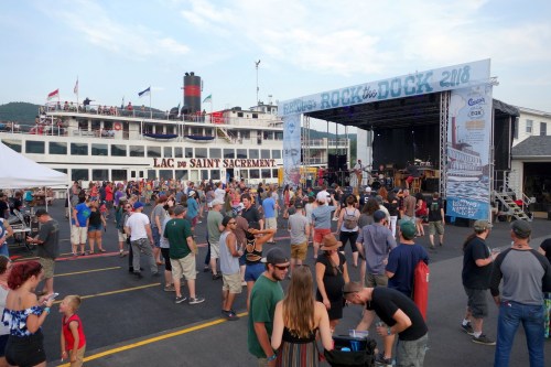 A lively outdoor concert scene with a large crowd, a stage, and a historic boat in the background.