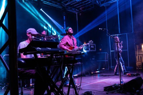 A band performs on stage with a keyboardist, guitarist, and vocalist under colorful lights.