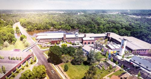Aerial view of a large industrial complex surrounded by greenery and a river, with roads and pathways visible.