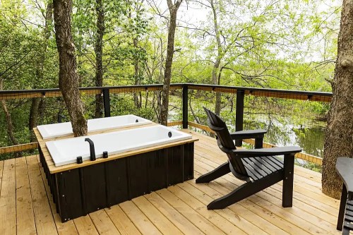A wooden deck with two hot tubs surrounded by trees and a view of a serene body of water. An Adirondack chair is nearby.