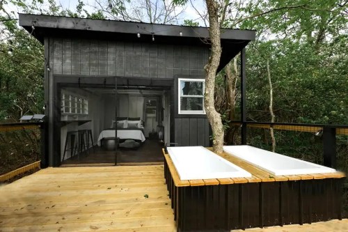 A modern cabin with large windows, surrounded by trees, featuring two outdoor bathtubs on a wooden deck.