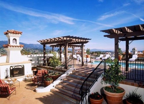 Rooftop terrace with seating, potted plants, and a pool, set against a clear blue sky and distant mountains.