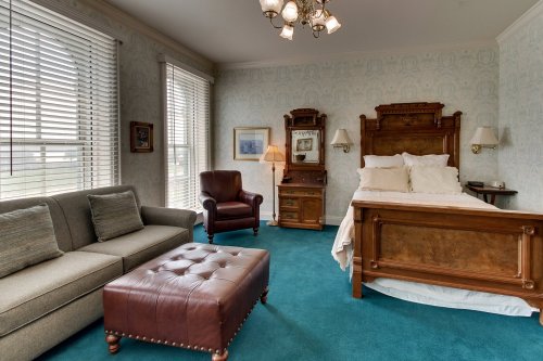 Cozy hotel room featuring a bed, sofa, armchair, and vintage decor with teal carpet and natural light from windows.