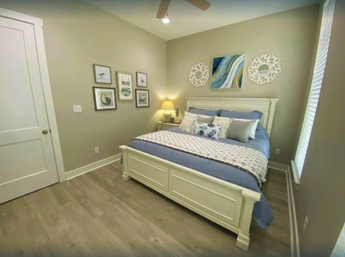 Cozy bedroom with a white bed, blue bedding, artwork on the walls, and natural light from a window.