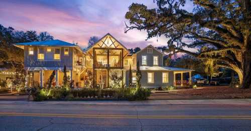 A beautifully lit two-story house at dusk, surrounded by trees and greenery, with a colorful sky in the background.