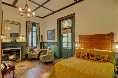 Cozy bedroom with a yellow quilt, fireplace, armchairs, and large windows, featuring vintage decor and warm lighting.
