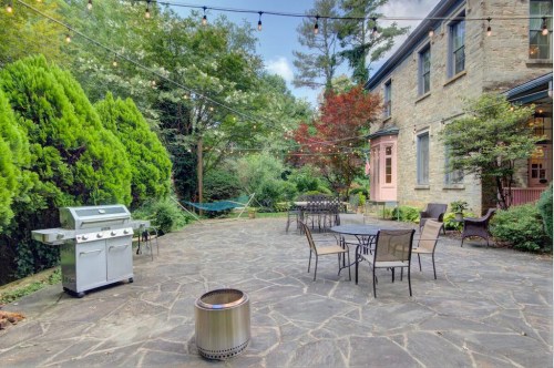 A stone patio with a grill, seating, and string lights, surrounded by greenery and a hammock in a serene outdoor setting.
