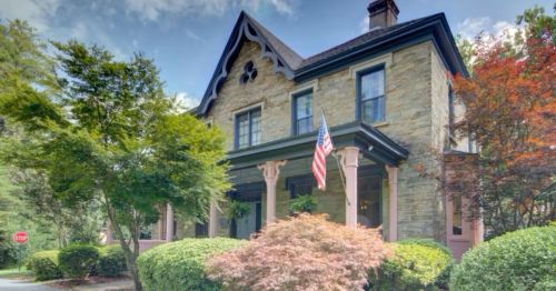 A charming stone house with a porch, American flag, and lush greenery in front, set against a clear blue sky.