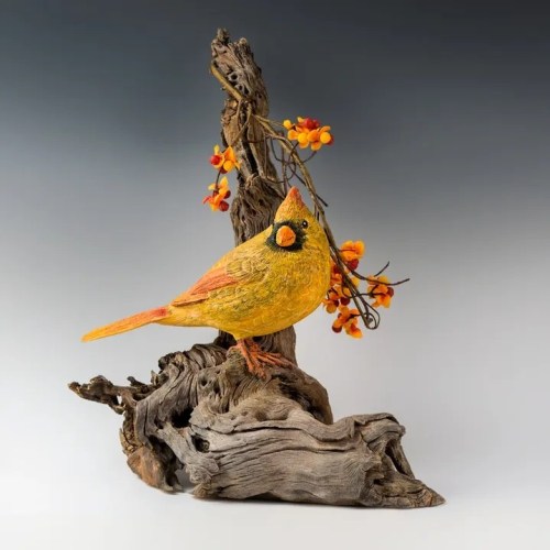 A colorful bird sculpture perched on a twisted wooden base, surrounded by orange flowers.