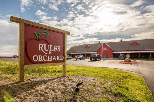 Sign for Rulfs Orchard in front of a red building, with cars parked and a cloudy sky in the background.