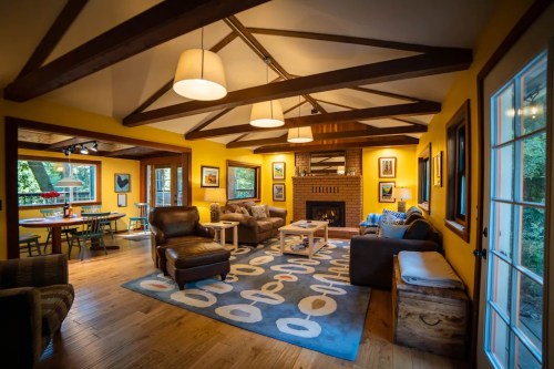 Cozy living room with yellow walls, wooden beams, comfortable furniture, and a fireplace, featuring a large area rug.