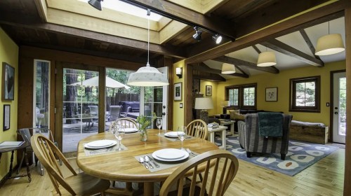 Cozy dining area with a wooden table, surrounded by yellow walls and large windows, leading to an outdoor deck.