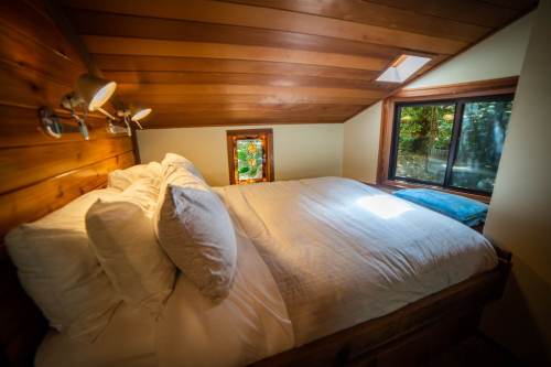 Cozy bedroom with a wooden ceiling, large bed, and windows showcasing greenery outside.