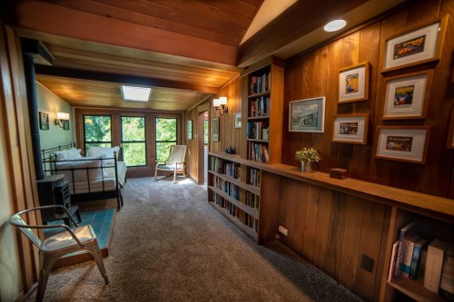 Cozy bedroom with wooden walls, a bookshelf, and a view of greenery through large windows. Soft carpet and natural light.