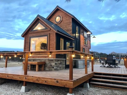 A cozy wooden cabin with a deck, surrounded by nature under a cloudy sky, featuring outdoor seating and warm lighting.