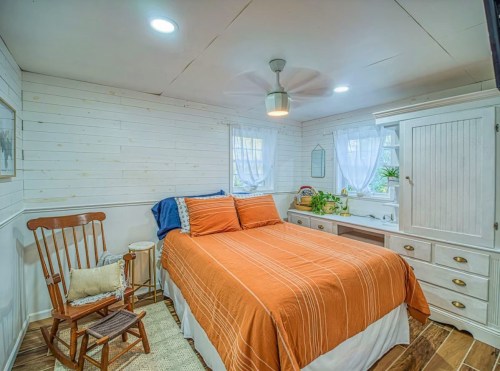 Cozy bedroom with a wooden rocking chair, orange bedspread, and natural light from windows. White walls and plants add warmth.