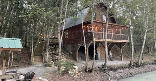 A rustic wooden cabin on stilts surrounded by trees, with a deck and string lights, near a calm waterway.