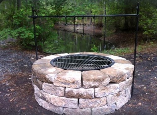 A stone fire pit with a metal grill and hooks, surrounded by trees and a calm waterway in the background.