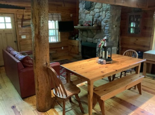 Cozy cabin interior featuring a wooden dining table, leather sofa, stone fireplace, and rustic decor.