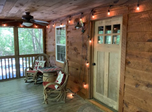 Cozy porch with wooden walls, string lights, a door, and two wicker chairs with decorative pillows.