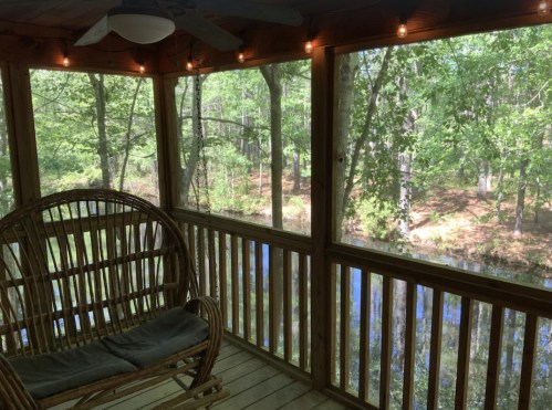 A cozy screened porch with a wicker chair, overlooking a serene wooded area and a calm pond.
