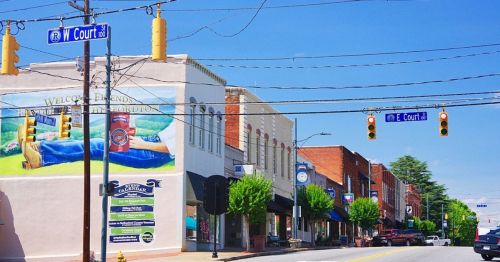 A sunny street view of a small town with shops, traffic lights, and a mural welcoming visitors.