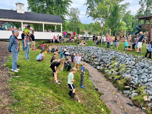 A crowd of people, including children, gather by a stream, watching colorful objects float by during a community event.