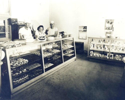 Black and white photo of three bakers in a bakery, standing behind a display case filled with various pastries and cakes.