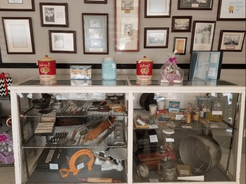 Display case filled with vintage kitchen tools, baking supplies, and framed historical photos on the wall.