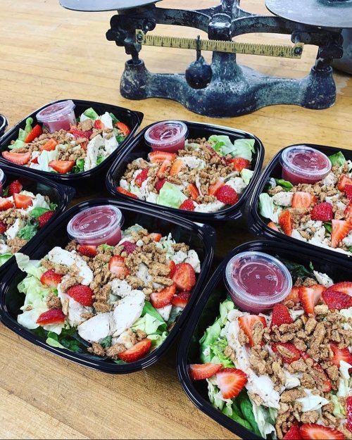 Six salad containers with chicken, strawberries, and crunchy toppings, placed near a vintage scale.