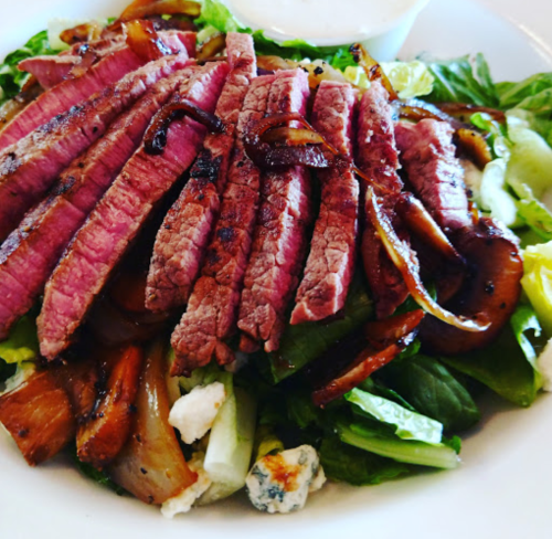 A fresh salad topped with sliced grilled steak, caramelized onions, and a side of dressing.