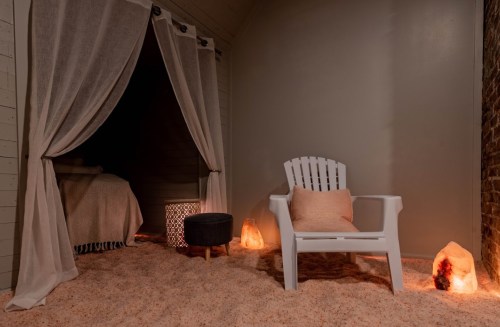 A cozy, softly lit room with a white chair, a small table, and salt lamps, featuring a sandy floor and draped curtains.