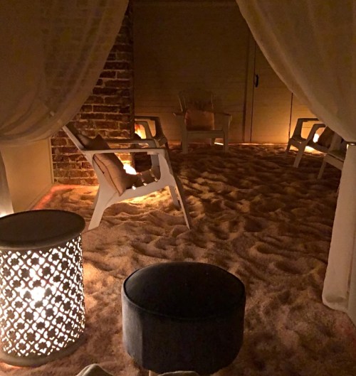 A cozy, dimly lit relaxation area with sandy flooring, chairs, and decorative lanterns.