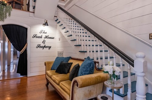 A cozy entryway featuring a yellow sofa, decorative flowers, and a staircase with a sign reading "The Oak House & Pantry."