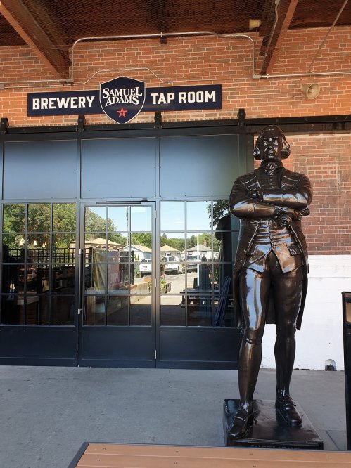 A bronze statue of a man stands outside the Samuel Adams Brewery Tap Room, with a sign above the entrance.