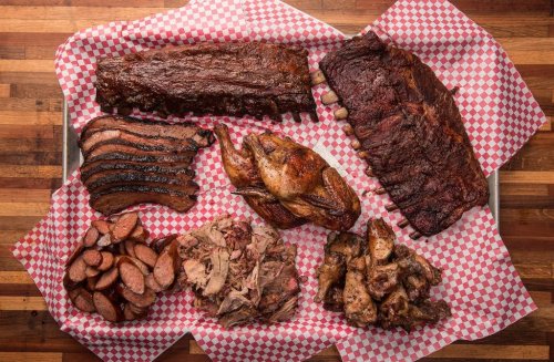 A platter of assorted barbecue meats including ribs, brisket, chicken, sausage, and pulled pork on checkered paper.