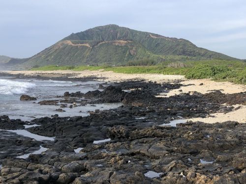 A rocky shoreline with a sandy beach and a green mountain in the background under a cloudy sky.