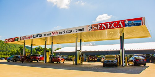 A truck stop with a large canopy, featuring the name "Seneca" and various vehicles parked outside under a blue sky.