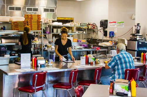A diner kitchen with two staff members working, one serving food and another preparing orders, with customers at the counter.