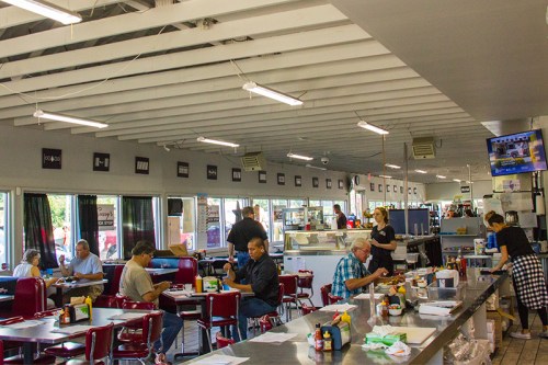 A busy diner interior with patrons eating, a server at the counter, and a TV displaying news in the background.