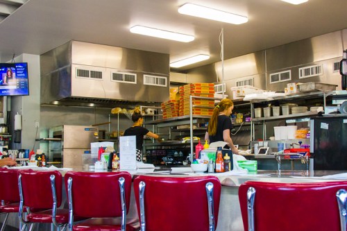 A diner kitchen with stainless steel appliances, two staff members working, and red chairs at the counter.