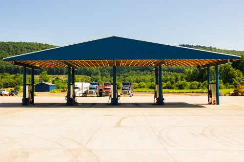 A large blue truck fueling station with multiple pumps under a roof, surrounded by green hills and clear skies.