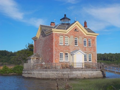 A historic brick lighthouse with a yellow trim, surrounded by water and greenery under a blue sky.