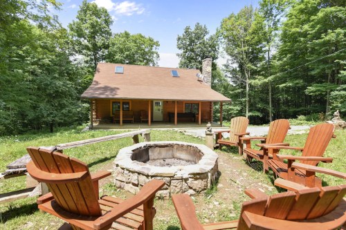 A cozy log cabin surrounded by trees, featuring a fire pit and wooden chairs in a serene outdoor setting.