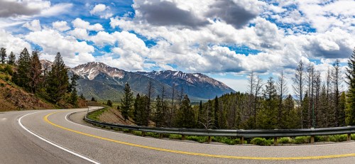 A winding road curves through a mountainous landscape under a blue sky with fluffy clouds and scattered trees.