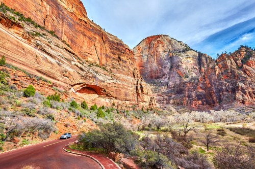 A scenic view of red rock cliffs and a winding road in a canyon, with sparse vegetation and a blue sky above.