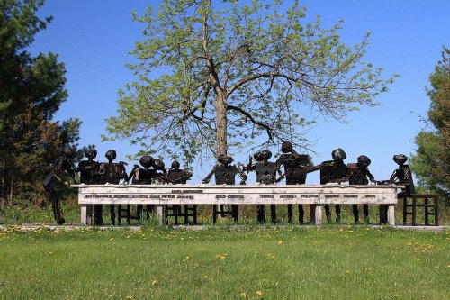 A group of metal sculptures depicting musicians seated at a bench under a tree, surrounded by green grass and blue sky.