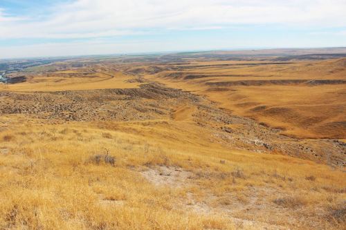 A vast, dry landscape with rolling hills and sparse vegetation under a blue sky with wispy clouds.