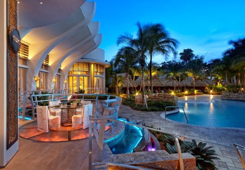 A modern outdoor dining area beside a pool, surrounded by palm trees and illuminated by soft evening lights.