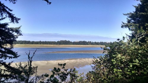 A serene landscape featuring a calm river, sandy banks, and trees under a clear blue sky.